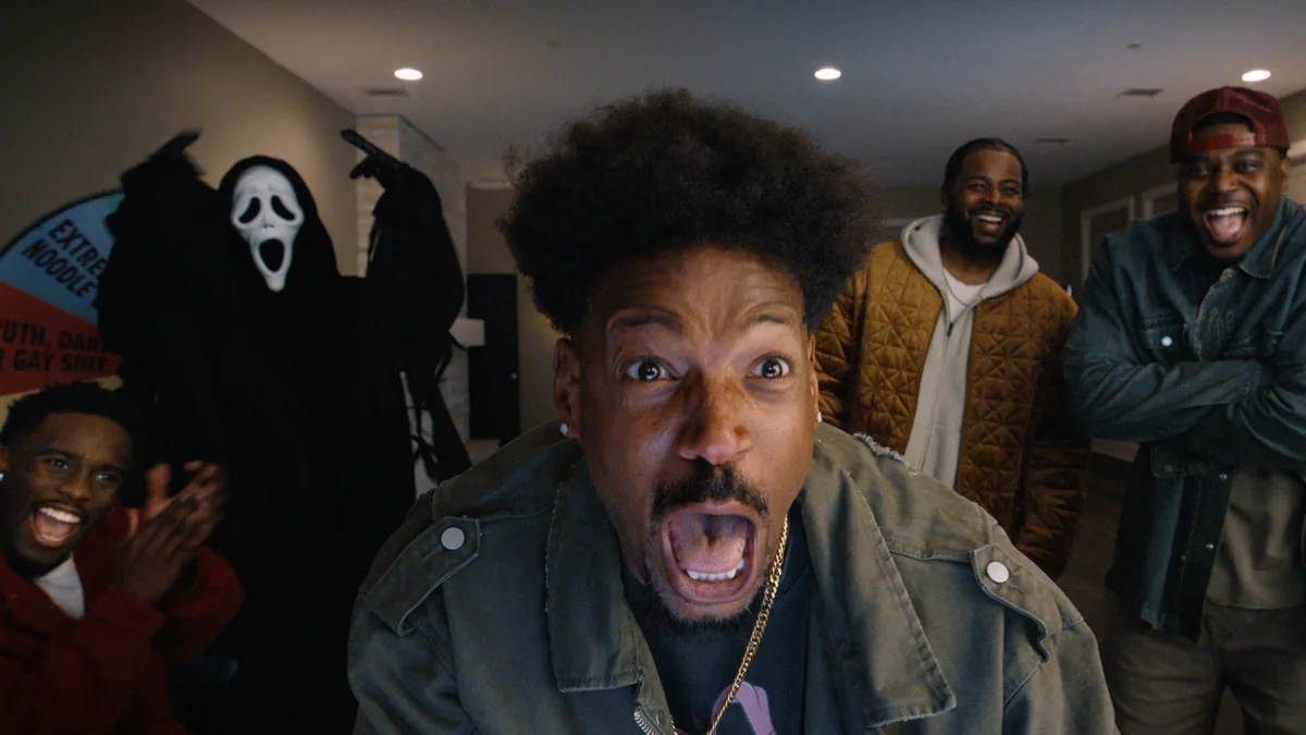 Scary Movie 2026 film sahnesi - Ghostface maskeli katil arka planda, onde korku icinde bagirak karakter ve gulen arkadaslari
