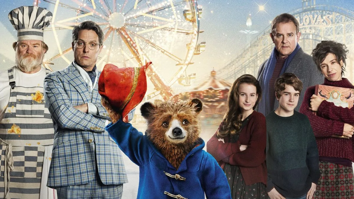 Paddington 2 filminde ayı Paddington, lunapark manzarası önünde şapkasını kaldırırken ailenin diğer üyeleriyle