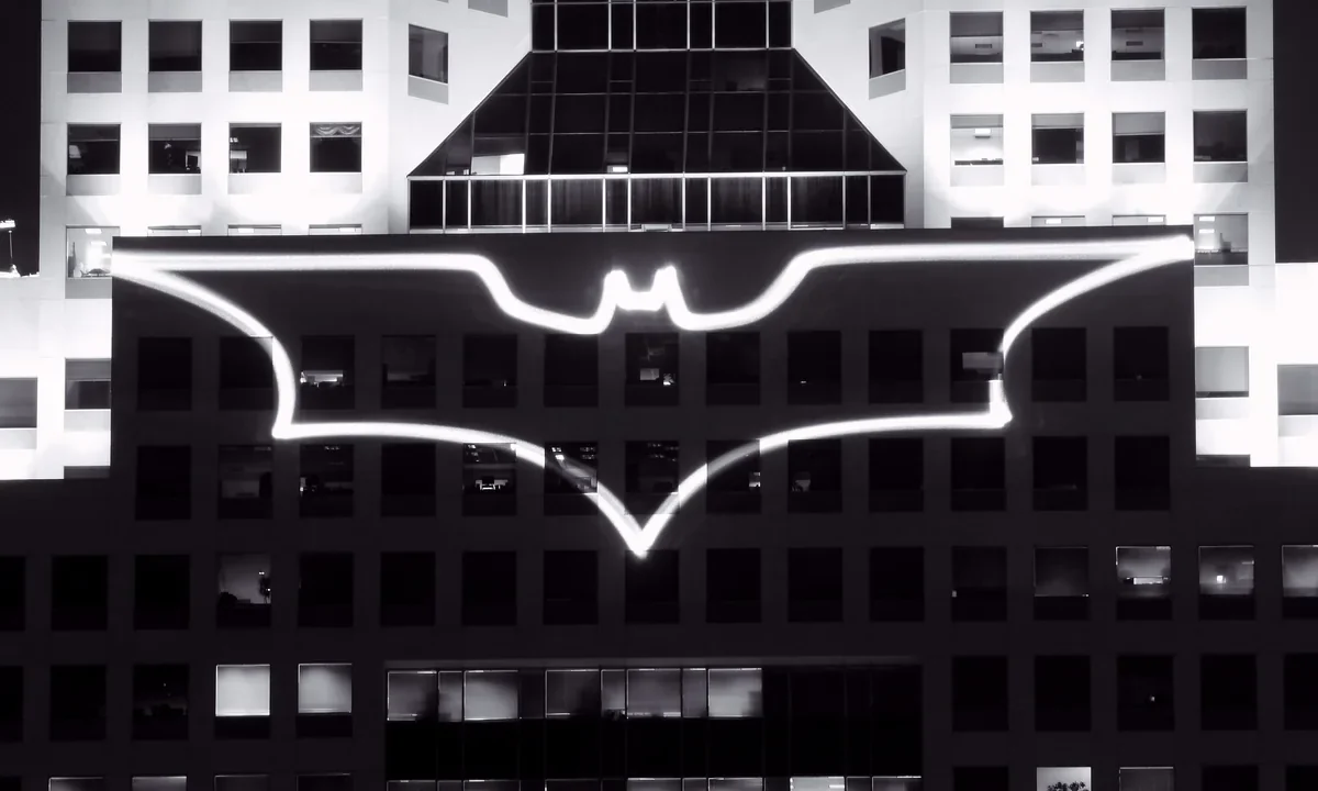 Batman'in yarasa logosunun bir gökdelen üzerine ışıkla yansıtıldığı gece görüntüsü, karanlık Gotham atmosferini simgeliyor