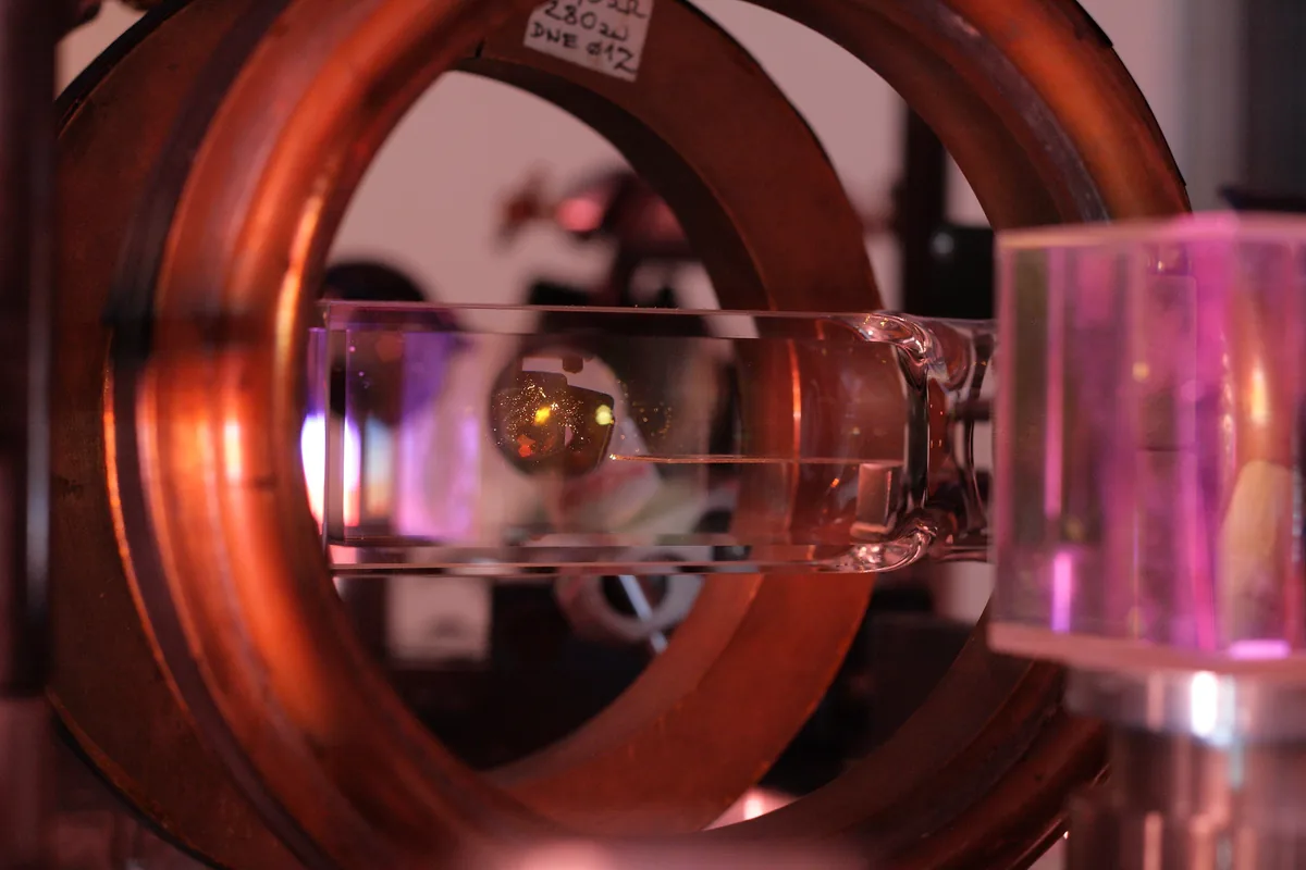 Magneto-optik tuzak (MOT) düzeneğinde vakum haznesi içinde parlayan turuncu-sarı soğuk rubidyum atom bulutu ve manyetik alan bobinleri